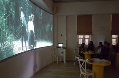 תמונה של אנשים צופים במיצג המרכזי בבית שיח נשים - סרט המוקרן על גבי מסך גדול על הקיר